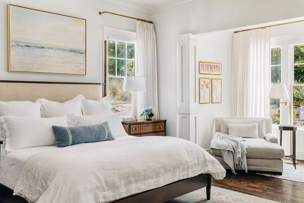 Bright, cozy bedroom with a king-sized bed, white bedding, decorative pillows, wooden nightstand with a lamp and plant, armchair with throw blanket, large windows with white curtains, and artwork on white walls.