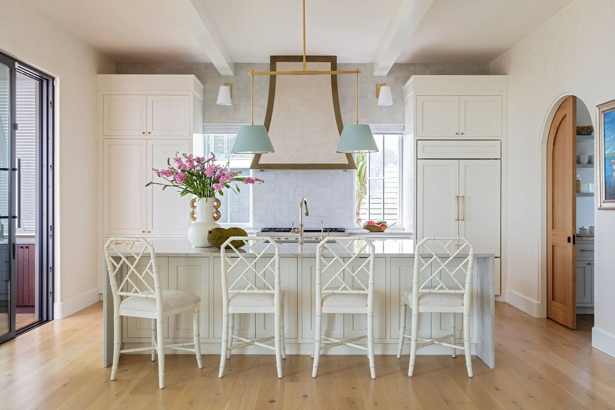 Modern white kitchen with a central island, four white chairs, large vase with pink flowers, two hanging pendant lights, and a white range hood.